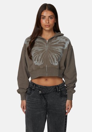 Przycięta brązowa bluza z kapturem z srebrnym motylem, zapinana z przodu na zamek, z długimi rękawami, zestawiona z wysokim stanem wyblakłych czarnych dżinsów.