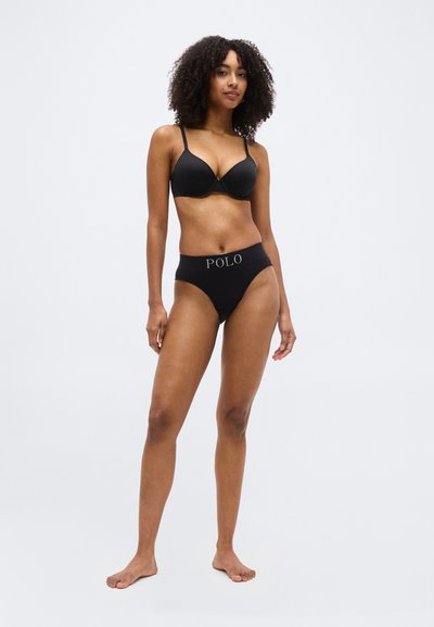 Γυναίκα με σγουρά μαλλιά που φοράει μαύρο σουτιέν και μαύρο εσώρουχο ψηλόμεσο με την ετικέτα «POLO», στεκόμενη ξυπόλητη σε απλό φόντο.