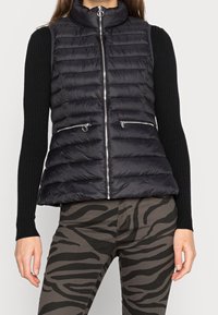 Frau trägt eine schwarze gesteppte Zip-Weste, ein schwarzes geripptes Langarmoberteil und graue Hosen mit schwarzen Zebra-Streifen.