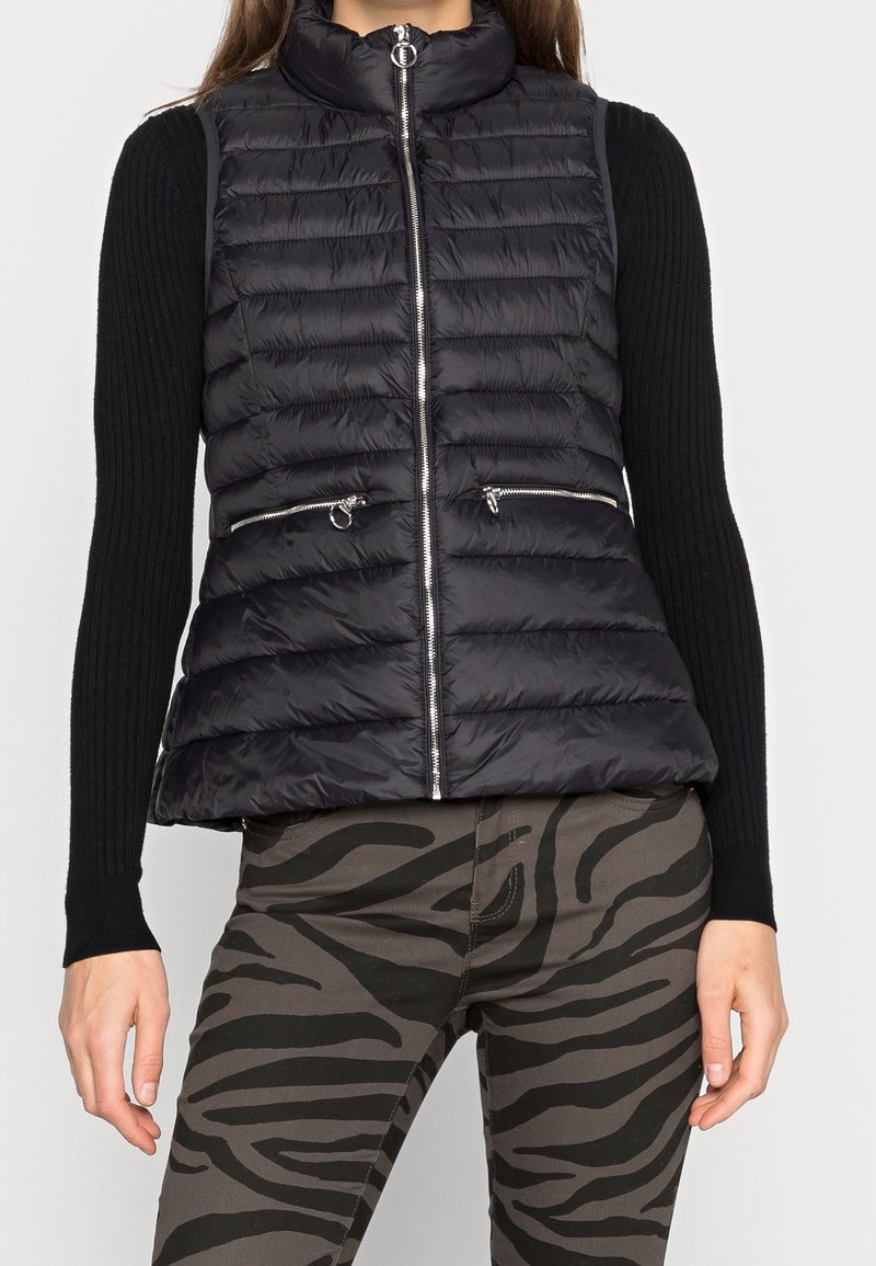 Frau trägt eine schwarze gesteppte Zip-Weste, ein schwarzes geripptes Langarmoberteil und graue Hosen mit schwarzen Zebra-Streifen.