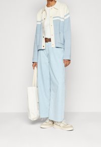 Jasnoniebieska dzianinowa kurtka z kremowymi akcentami, biała koszulka, jasne dżinsowe spodnie o szerokich nogawkach, beżowe trampki i biała torba na ramię.