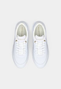 O pereche de adidași albi Tommy Hilfiger cu șireturi albe și cuiele aurii, prezentată dintr-o perspectivă de sus pe un fundal deschis.
