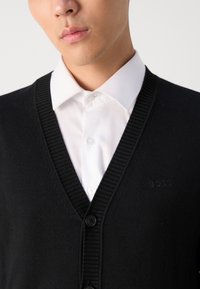 Cárdigan negro de punto con escote en V, bordes acanalados y cuatro botones negros. Lleva encima de una camisa blanca con cuello, que presenta un logotipo en relieve.