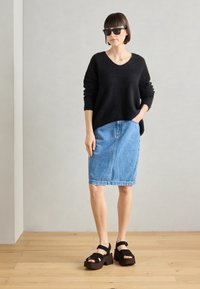 Een vrouw in een zwarte trui, een spijkerrok tot de knie, zwarte platform sandalen en een zonnebril, staand op een houten vloer tegen een eenvoudige muur.