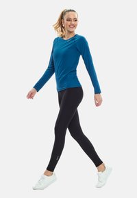 Młoda kobieta idąca, ubrana w niebieską bluzkę z długim rękawem, czarne legginsy i białe tenisówki na jednolitym białym tle.