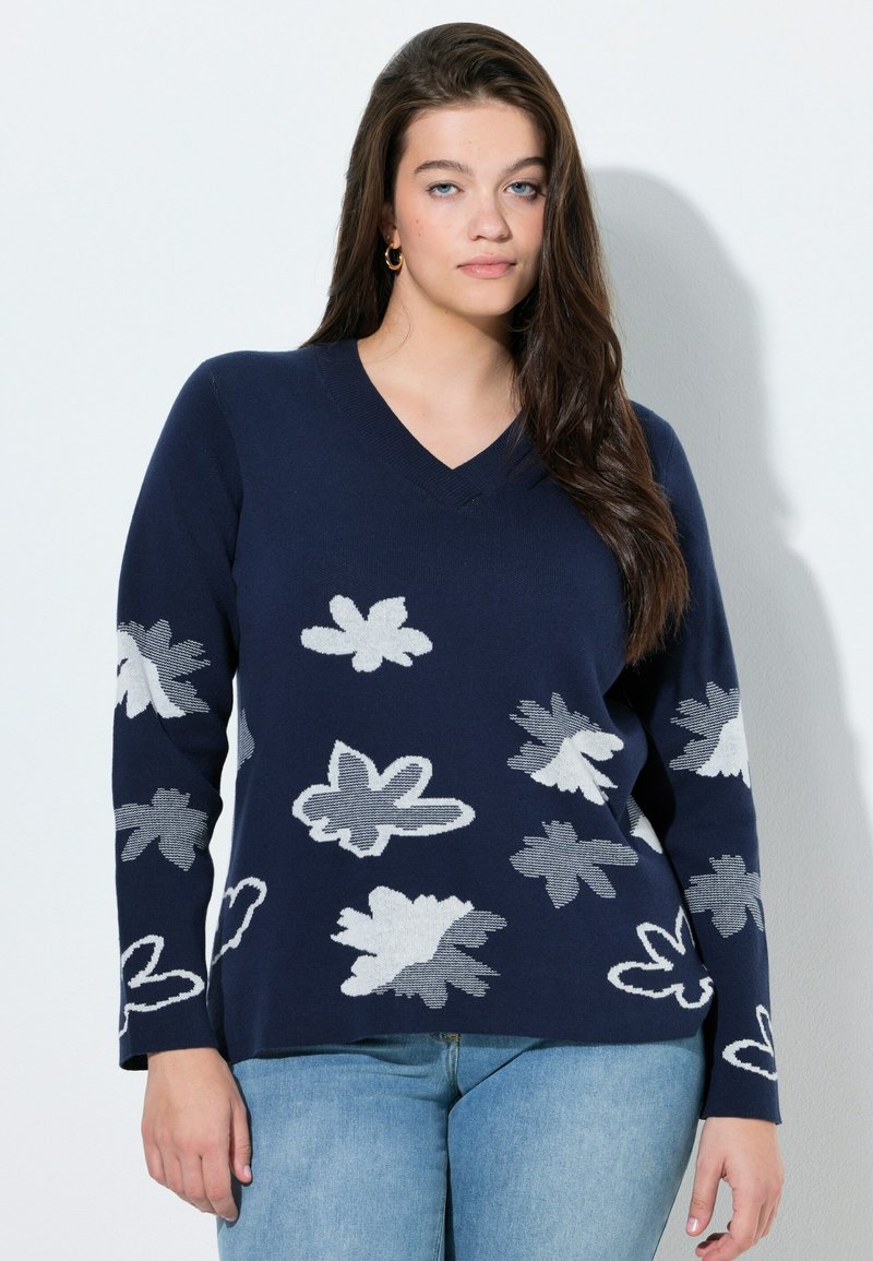 Granatowy sweter z dekoltem w serek, zdobiony abstrakcyjnymi białymi kwiatowymi wzorami, z długimi rękawami i miękką fakturą, zestawiony z jasnoniebieskimi dżinsami.