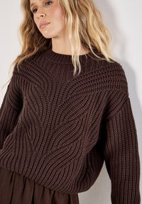 Brauner, gestrickter Pullover mit strukturierten Wellenmustern, weiten Ärmeln und rundem Ausschnitt. Der Stoff wirkt weich und dick.