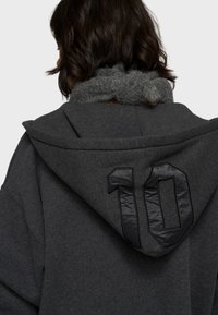 Grijze hoodie met een grote capuchon en een pluizige kraag. Heeft een zwart nummer "10" geborduurd op de achterkant. De stof lijkt zacht met een gestructureerde afwerking.