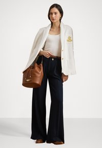Bílý blazer se zlatými akcenty, černé široké kalhoty, béžový top na ramínka, hnědá kožená taška na šňůrku a hnědé boty.