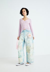 Lichtpaarse gebreide cardigan met gekartelde randen, gecombineerd met oversized denim jeans in patchworkstijl met verschillende pastelpatronen en texturen.