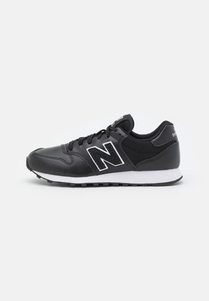Sort New Balance sneaker med hvid sål og stort N-logo med hvid kant på siden, vist i profil på en ensfarvet baggrund.