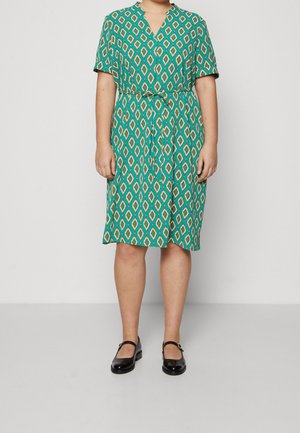 Woman wearing a green patterned knee-length dress with a tie waist and black Mary Jane shoes standing against a plain background.
