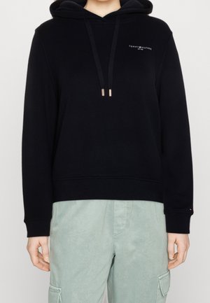 Czarna bluza z kapturem Tommy Hilfiger z troczkami, noszona z jasnobarwnymi zielonymi spodniami typu cargo, model pokazany od szyi do bioder na białym tle.