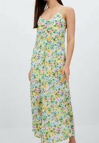 Robe longue fleurie avec des fines bretelles, fabriquée en tissu léger. Présente un fond jaune avec des motifs floraux roses, bleus et verts.