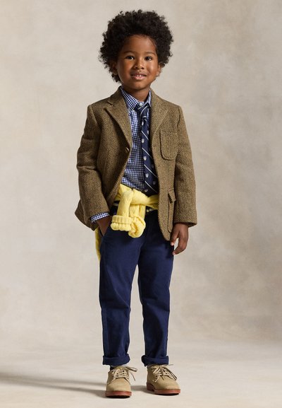 Niño joven que lleva una chaqueta marrón, camisa azul a cuadros con corbata, pantalones azul marino, zapatos beige y un jersey amarillo atado alrededor de su cintura.