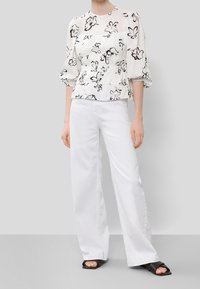 Weißes Blumenshirt mit schwarzen Umrandungen, Puffärmeln und geraffter Taille, kombiniert mit weißen weit geschnittenen Hosen und schwarzen Rutschen-Sandalen.