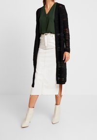 Cárdigan negro de punto sobre una blusa verde, combinado con una falda de mezclilla blanca con aberturas. Botines blancos con punteras puntiagudas completan el conjunto.