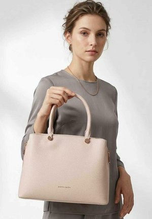 Frau mit hellbraunen Haaren in einem grauen Oberteil hält eine große hellrosa Lederhandtasche mit zwei Henkeln und goldener Hardware.