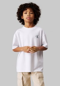 Camiseta de algodón blanca con cuello redondo, mangas cortas y un pequeño logotipo oscuro en el lado izquierdo del pecho. Combinada con pantalones cargo beige.