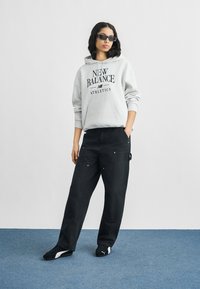 Γυναίκα που φοράει μαύρα γυαλιά ηλίου, γκρι φούτερ New Balance, φαρδιά μαύρη παντελόνια και μαύρα παπούτσια Puma, στέκεται σε μπλε χαλί.