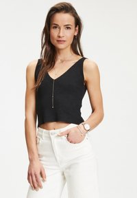 Top corto nero a costine con scollatura a V, abbinato a jeans bianchi a vita alta. Gli accessori includono un orologio e collane a strati.