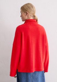 Rode trui met een hoge kraag, losse pasvorm en lange mouwen. De stof lijkt zacht en glad. Draagt over een denim rok.