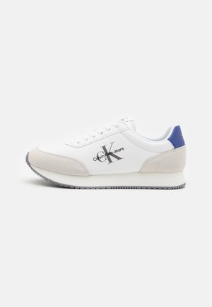 Zapatilla blanca con parte delantera de ante gris, trabilla trasera azul y logo de Calvin Klein Jeans en gris. Cierre de cordones planos, suela de goma y interior acolchado.