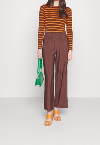 Gestreepte lange mouwen top in oranje en bruin, bruine wijde broek, groene handtas en oranje en beige blokhak sandalen.