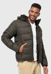 Grüne gepolsterte Jacke mit Reißverschluss und Kapuze. Verfügt über zwei Seitentaschen und strukturierte Stoffoberfläche. Getragen über einem weißen Hemd.