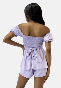 Lila Off-Shoulder-Top mit gerafftem Oberteil, Rüschensleeves und einem gebundenen Gürtel in der Taille, kombiniert mit passenden Shorts. Weicher Stoff.