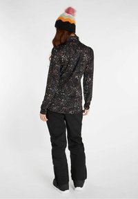 Maglietta nera a maniche lunghe con un motivo maculato in vari colori, abbinata a pantaloni neri e un berretto di lana multicolore con un pom-pom rosa.