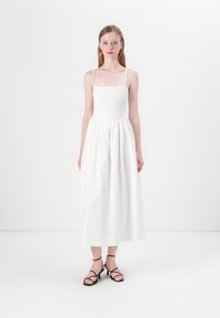 White sleeveless dress with a fitted smocked bodice and a flowing, gathered skirt. Paired with strappy black sandals. Simple and minimal design.