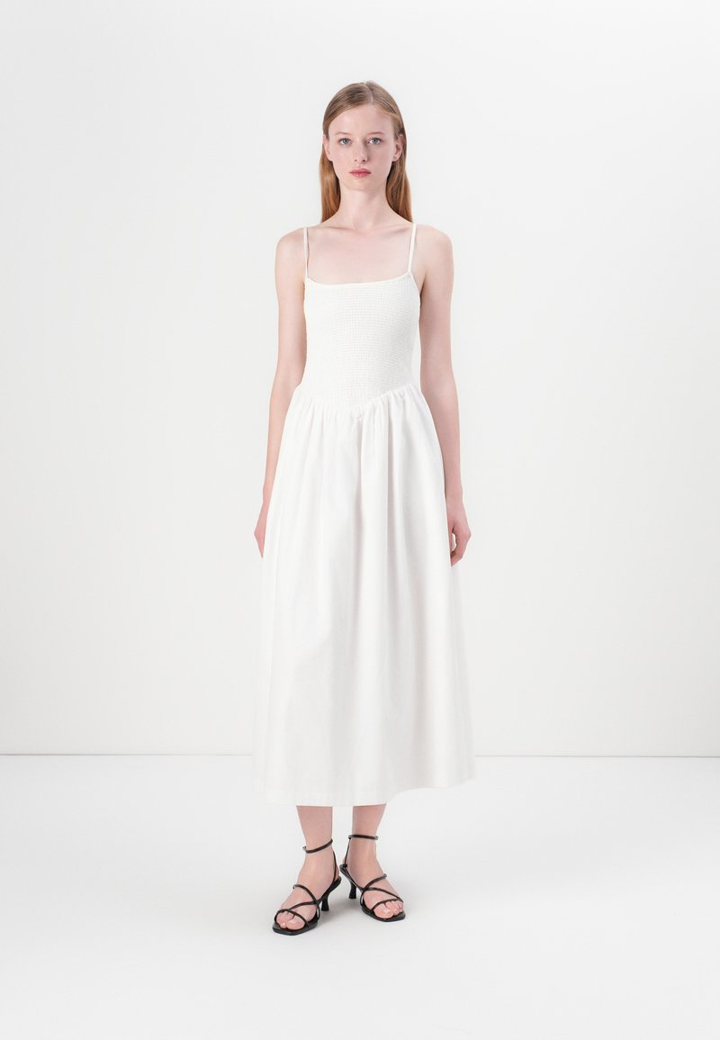 Vitt ärmlöst klänning med en figurnära smockad bustier och en flödande, insamlad kjol. Kombinerad med svarta sandaler med remmar. Enkel och minimalistisk design.