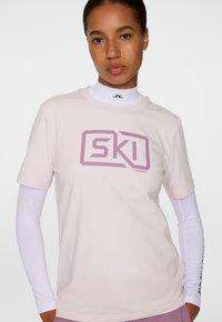 Jasnoróżowa bawełniana koszulka z wyraźnym fioletowym nadrukiem "SKI" w geometrycznym stylu, noszona z białą podkoszulką z długim rękawem pod spodem.