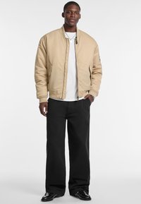 Beige bomber jacket with a front zipper, ribbed cuffs, and pockets, worn over a white t-shirt and paired with black, loose-fit pants.