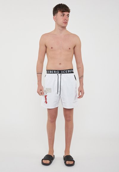 Costumi da bagno bianchi con cordoncino e vita neri, caratterizzati da un design grafico rosso e una piccola etichetta. Consegna con ciabatte nere.