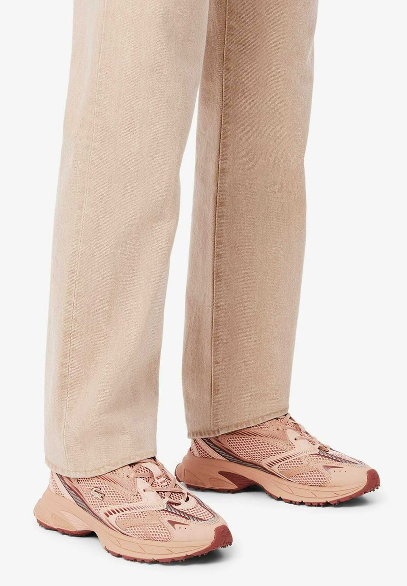 Beige Hosen mit weitem Bein, kombiniert mit pink-braunen Mesh-Sneakern, stehend auf einer weißen Oberfläche.