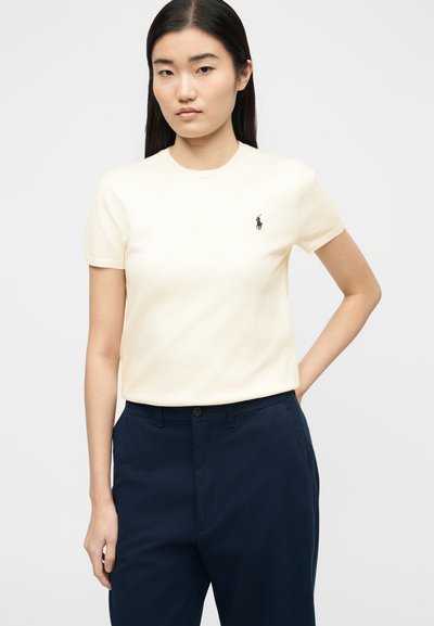 Νεαρή γυναίκα που φοράει κρεμ πλεκτό τοπ με κοντά μανίκια και μικρό κεντημένο λογότυπο, καθώς και μπλε σκούρο παντελόνι, στέκεται με το ένα χέρι στη μέση.