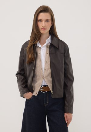 Joven mujer con cabello largo y castaño lleva chaqueta de cuero marrón oscuro, cárdigan beige, camisa blanca, cinturón marrón y vaqueros anchos azul oscuro.