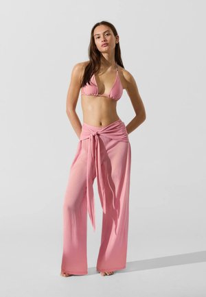 Donna che indossa un top bikini rosa con pantaloni ampi trasparenti abbinati legati in vita, in piedi scalza su sfondo neutro.