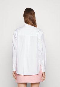 Camicia bianca a maniche lunghe con piega sul retro, polsini a bottoni e orlo curvo, indossata sopra una gonna rosa strutturata. Tessuto liscio, design classico.