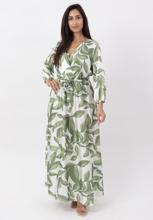 Femme portant une robe longue blanche à imprimé feuilles vertes, manches longues et ceinture à la taille, debout avec une main sur la hanche.
