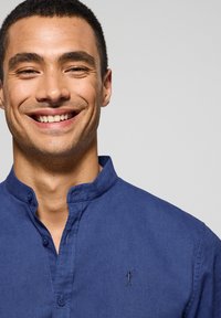 Niebieska koszula z kołnierzykiem, zapinana na guziki, wykonana z gładkiego materiału. Posiada subtelną haftowaną aplikację po lewej stronie klatki piersiowej.