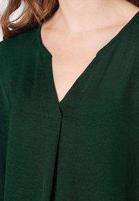 Närbild av en person som bär en mörkgrön t-shirt med V-ringning, med långt brunt hår synligt på axeln.