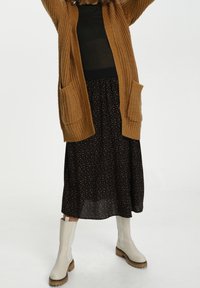 Brązowy dziergany kardigan z naszywanymi kieszeniami, zestawiony z czarną spódnicą w pomarańczowe kropki, uzupełniony białymi kozaczkami za kolano.