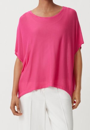 Helles pinkes, übergroßes Blusenshirt aus transparenter Stoff, mit einem runden Halsausschnitt und kurzen Ärmeln, kombiniert mit leichten weißen Hosen.