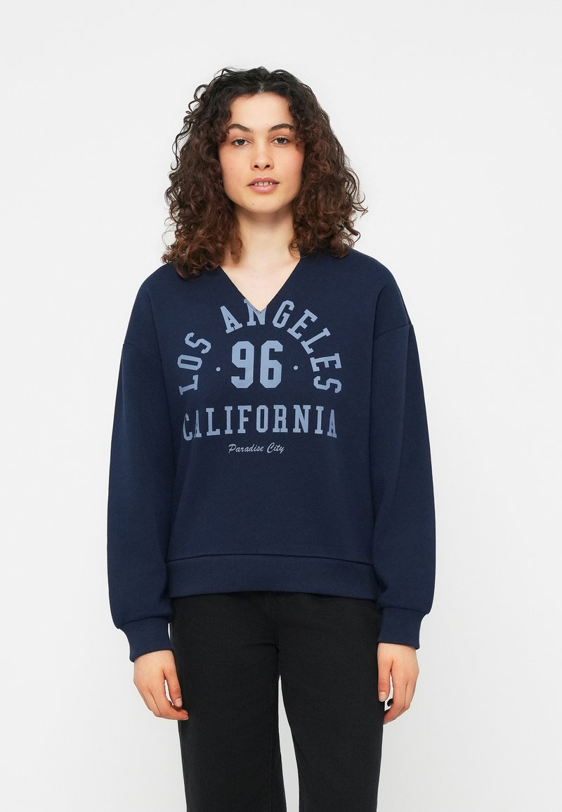 Jeune femme aux cheveux bouclés portant un sweat-shirt bleu marine avec le texte « Los Angeles 96 California Paradise City », debout devant un fond uni.