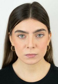Ritratto di una persona con lunghi capelli castani e occhi verdi. Indossa piccoli orecchini a cerchio d'oro e una maglia nera. Lo sfondo è neutro.