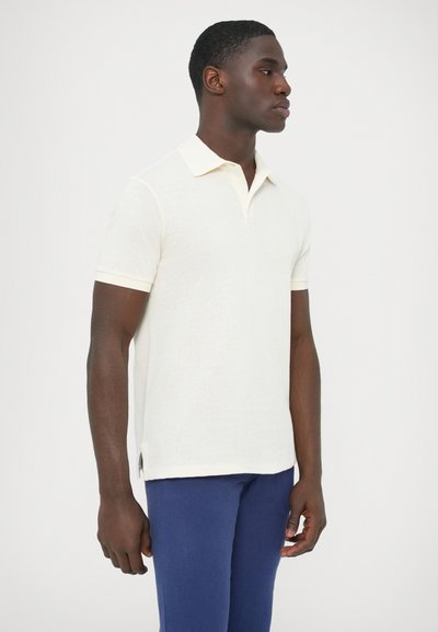 Polo tričko s krátkym rukávom z pletenej látky v smotanovej farbe, s golierom, kontrastným bielym lemovaním a bočnými rozparkami pre ľahší pohyb.