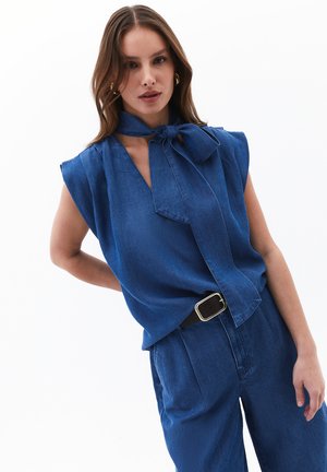 Blå denim top med v-hals og sløjfedetalje ved halsudskæringen, med brede ærmer og en glat tekstur. Sat sammen med skræddersyede bukser.